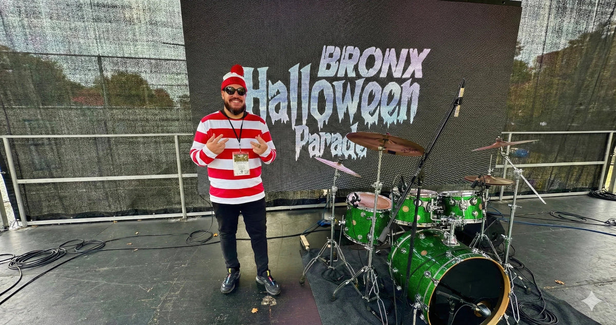 Man in red and white striped shirt and hat standing next to green drum set on stage at Bronx Halloween Parade