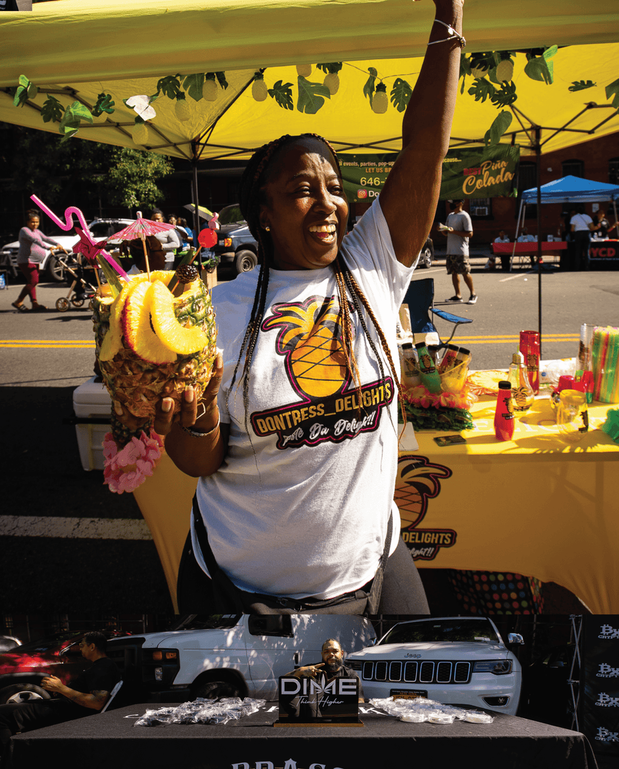 Person holding a pineapple drink at an outdoor event with a yellow canopy and various items on display.