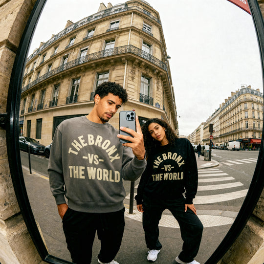 Two people wearing 'The Bronx vs The World' sweatshirts taking a selfie in a city street.