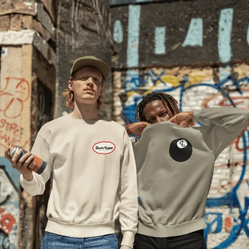Two people wearing sweatshirts with logos in front of a graffiti-covered wall.